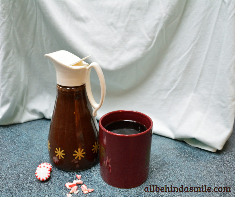An antique syrup bottle full of peppermint mocha syrup beside a reddish coffee mug