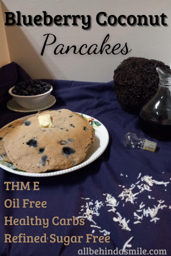bluish purple surface with a plate of blueberry coconut pancakes, shredded coconut, a bowl of fresh blueberries, and syrup in front of a coconut on it
