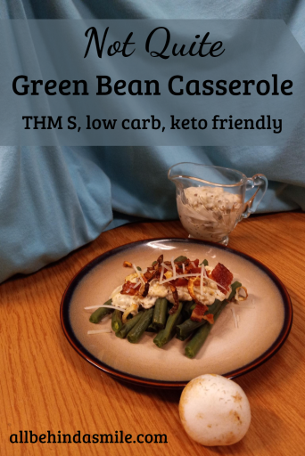 A tan plate with green beans topped with white sauce, bacon, cheese; a small crystal gravy dish with white sauce in the back right and a white mushroom in the front with a blue background and wood table: text is Not Quite Green Bean Casserole, THM S, low carb, keto friendly