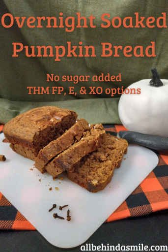 An orange and black checkered cloth under a white cutting board holding a sliced loaf of overnight soaked pumpkin bread with a knife beside it and a white pumpkin in front of the green cloth background