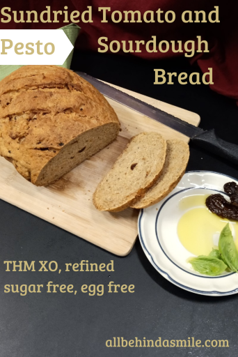 Text: Sundried Tomato and Pesto Sourdough Bread THM XO, refined sugar free, egg free Image: a loaf of sourdough with a small plate of oil, basil leaves, and sundried tomatoes